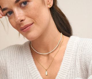 Eine Frau trägt zwei Ketten: eine Kette aus 925er Silber vergoldet, mit Perlmutt und eine Kette aus 925er Silber vergoldet, mit Süßwasserzuchtperlen. Sie trägt Ohrstecker aus 925 Silber vergoldet, mit Sonnenschliff.