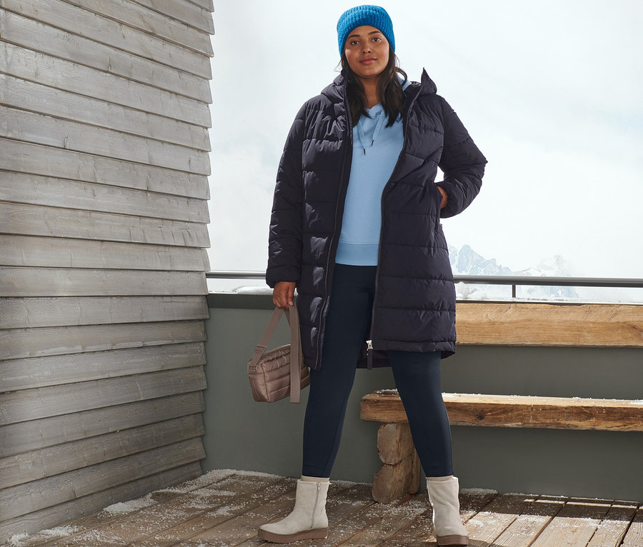 Eine Frau posiert draußen auf einer Holzterrasse mit Steppmantel in Dunkelblau, Strickmütze in Blau, Kapuzen-Sweatpullover, Thermotight in Blau, Veloursleder-Boots und Gesteppte Umhängetasche.
