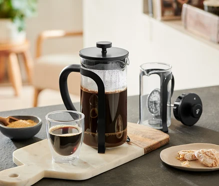 Eine schwarze Siebstempelkanne mit 800 ml Fassungsvermögen für 6 Tassen steht neben einem Glas Kaffee auf einem Marmorbrett.