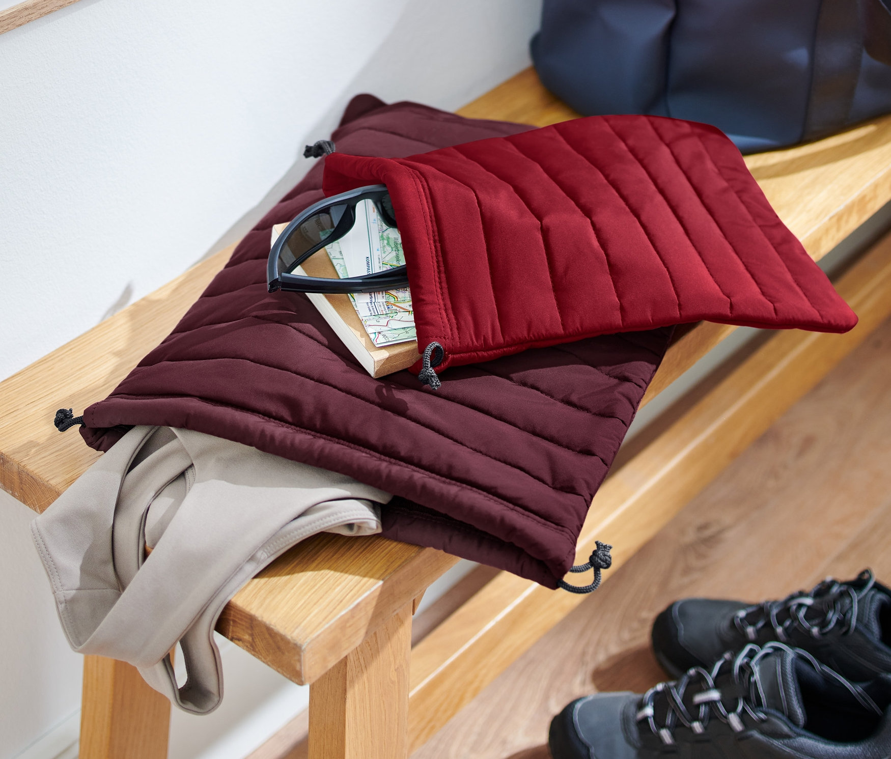 Auf einer Bank liegen 2 gesteppte Organizer, einer bordeauxrot und einer weinrot, mit Inhalt. Schuhe stehen auf dem Boden.