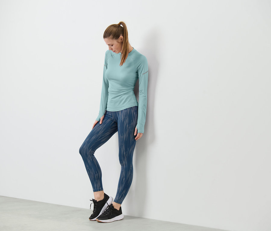 Eine Frau lehnt an einer Wand und trägt eine Sporttight, silberfarben reflektierend, und ein langärmliges Oberteil.