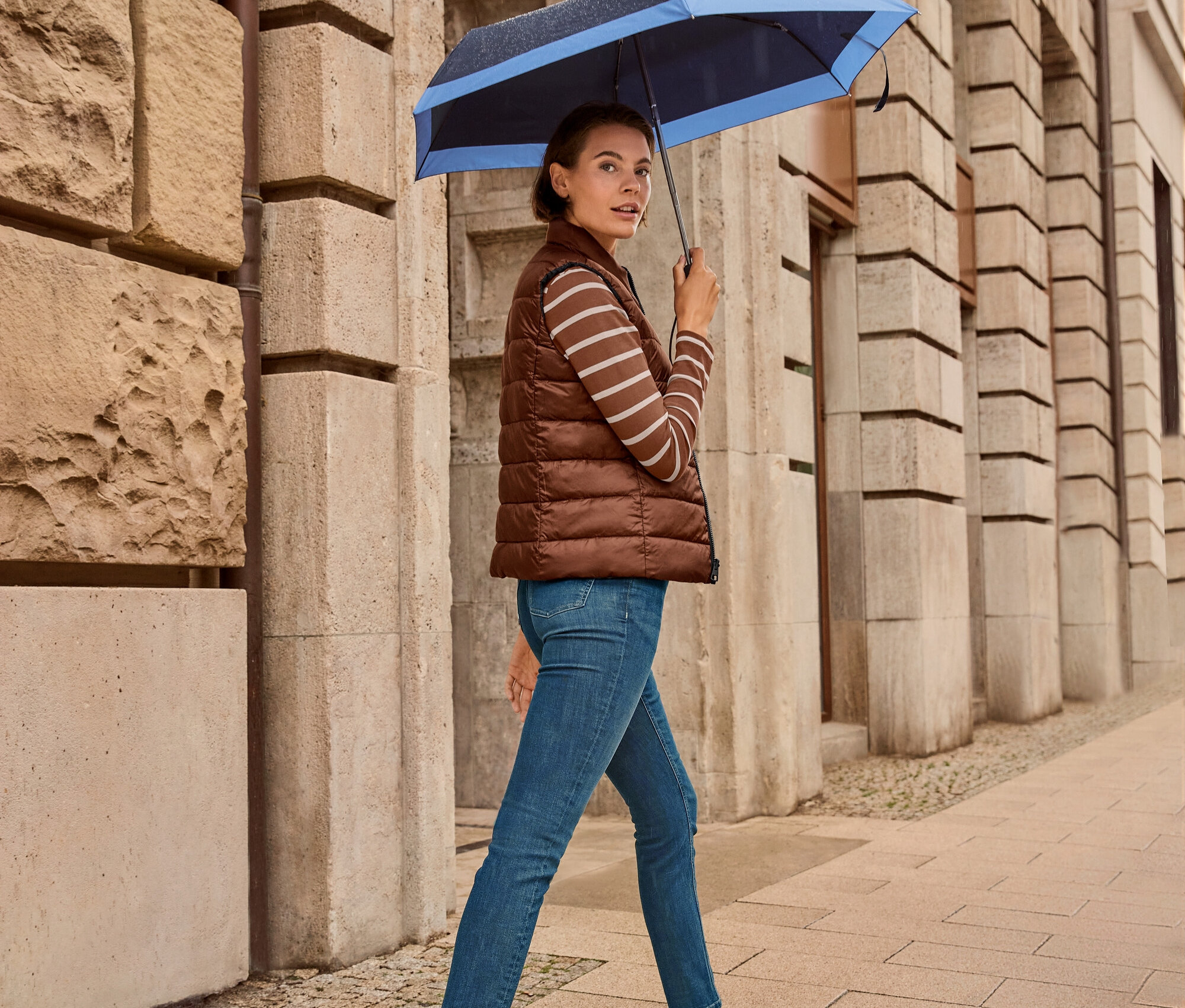Eine Frau geht mit einem Regenschirm auf dem Gehweg, sie trägt eine Wende-Steppweste, Slim Jeans – Fit »Emma«, dark blue und Leder-Sneaker.