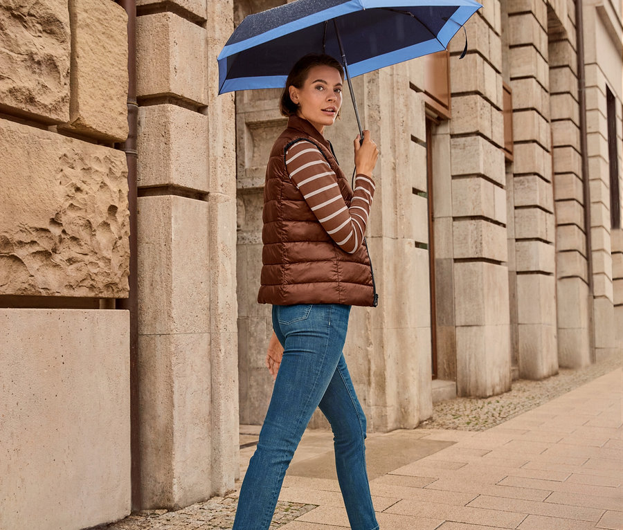Eine Frau geht mit einem Regenschirm auf dem Gehweg, sie trägt eine Wende-Steppweste, Slim Jeans – Fit »Emma«, dark blue und Leder-Sneaker.