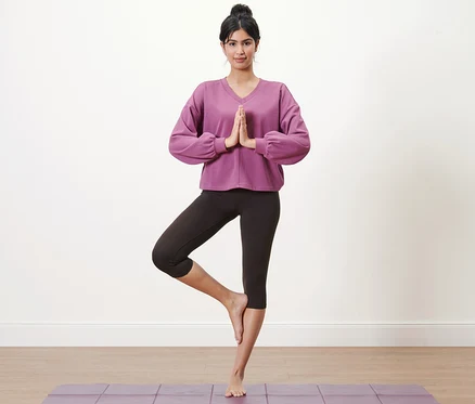 Frau steht in der Baumposition auf einer lila Yogamatte und trägt eine 3/4-Bodyforming-Sporttight.