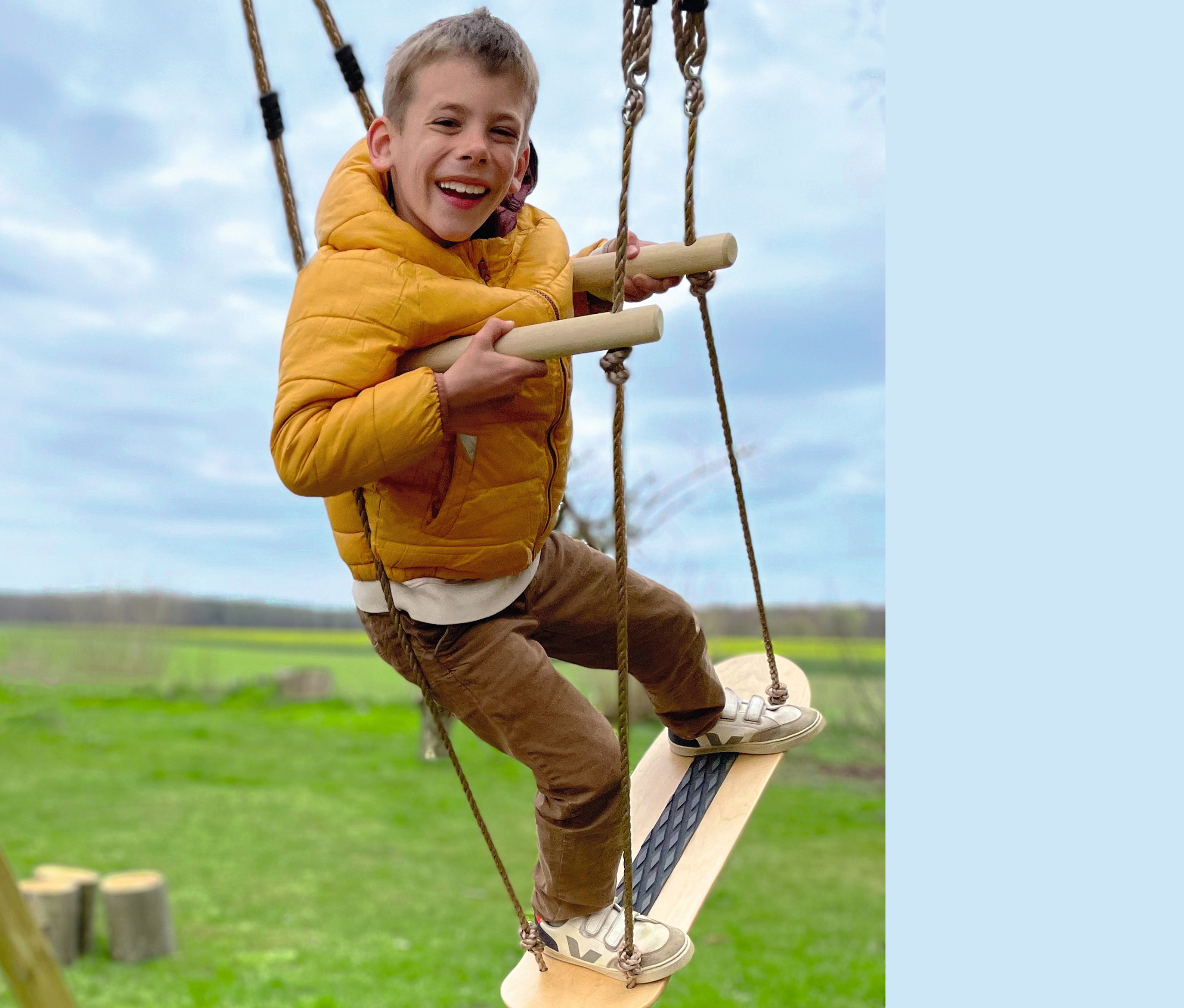 Ein Junge in gelber Jacke lacht und spielt auf einer small foot Skateboard-Schaukel.
