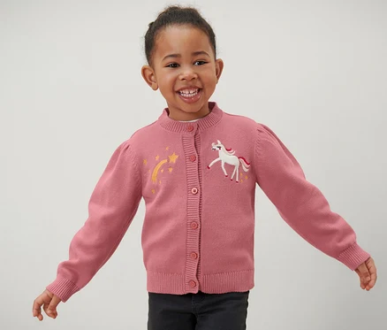 Lächelndes Mädchen trägt eine rosa Kinder-Strickjacke mit Einhorn- und Stern-Applikationen.