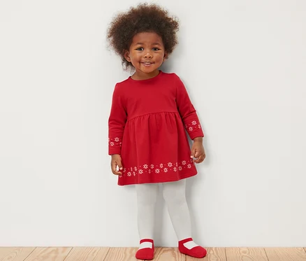 Ein lächelndes kleines Mädchen in einem roten Kleid mit Schneeflocken steht auf einem Holzboden und trägt weiße Strumpfhosen und rote Schuhe. Sie trägt auch 2 Baby-Thermostrumpfhosen, Ballerina.