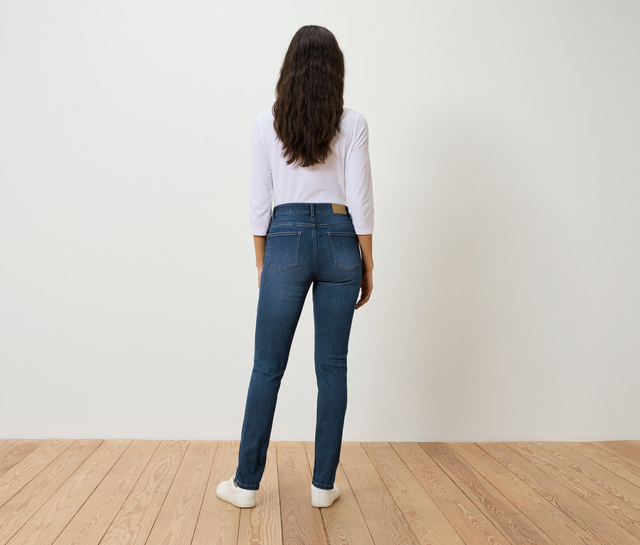 Eine Frau steht mit dem Rücken zur Kamera und trägt blaue Jeans, ein weißes Dreiviertelarm-Shirt und weiße Turnschuhe.