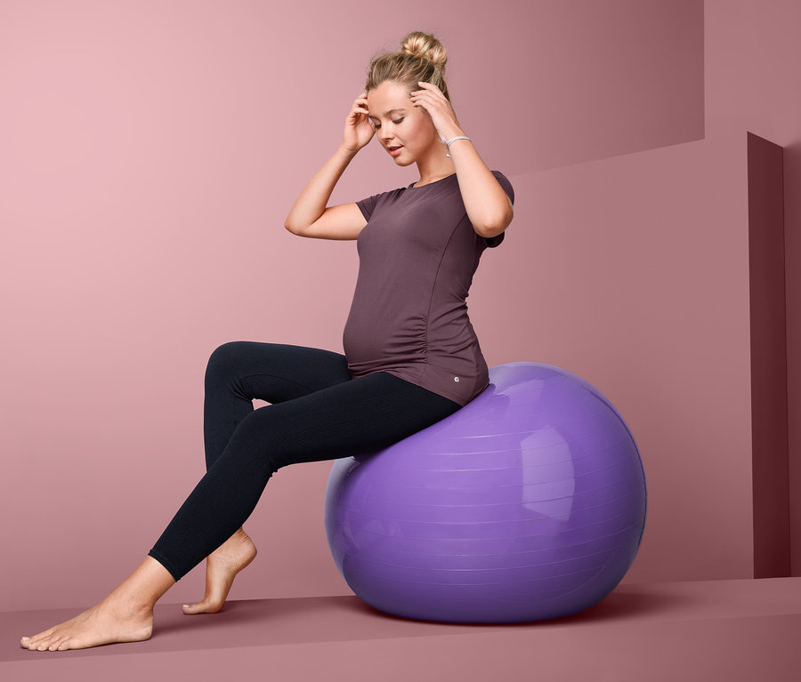 Schwangere Frau sitzt auf lila Gymnastikball, trägt ein Umstands-Sportshirt und schwarze Leggings.