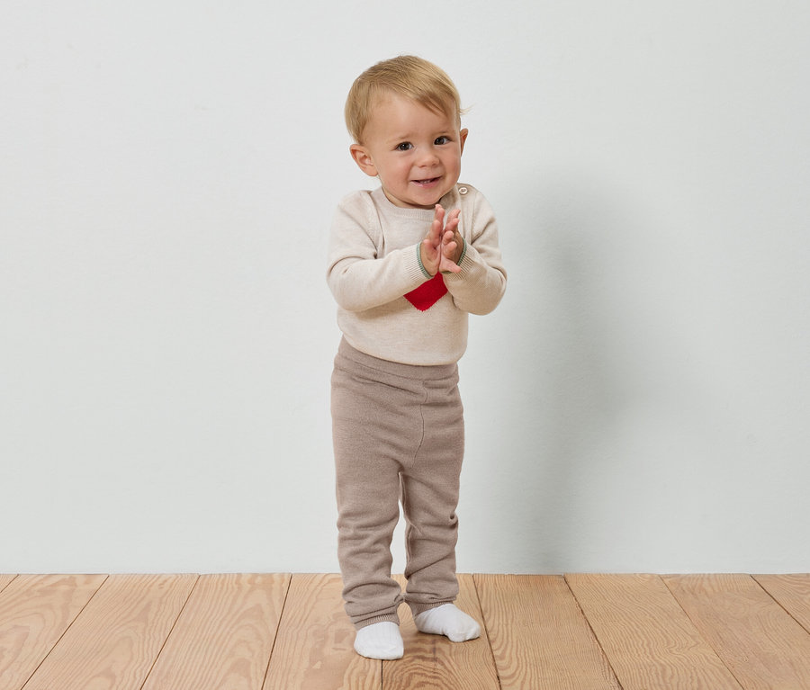 Ein Baby mit blonden Haaren klatscht in die Hände auf einem Holzboden vor einer weißen Wand.