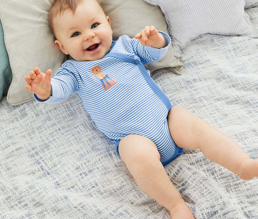 Lächelndes Baby liegt auf dem Rücken auf einem Bett in einem blau-weiß gestreiften Langarmbody. Das Baby trägt einen 3 Langarm-Bodys.