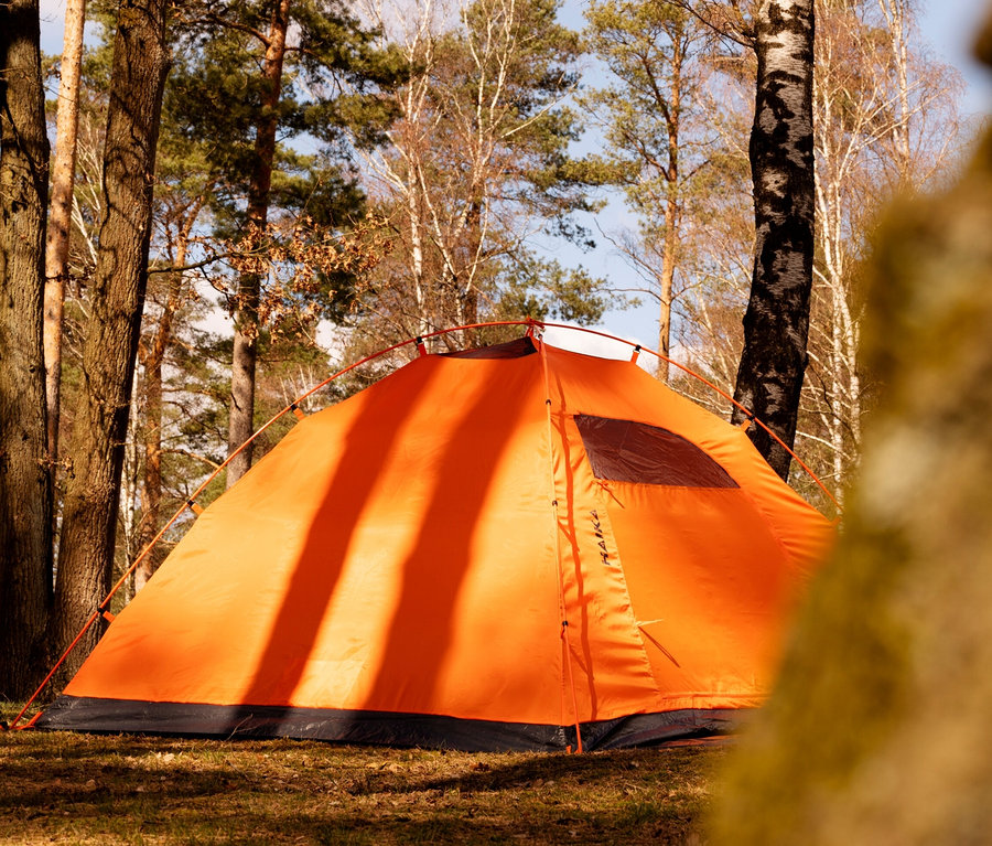 Ein orangefarbenes Tambu 2-Personen-Kuppelzelt »HAIKA 2» steht auf einer Wiese zwischen Bäumen.