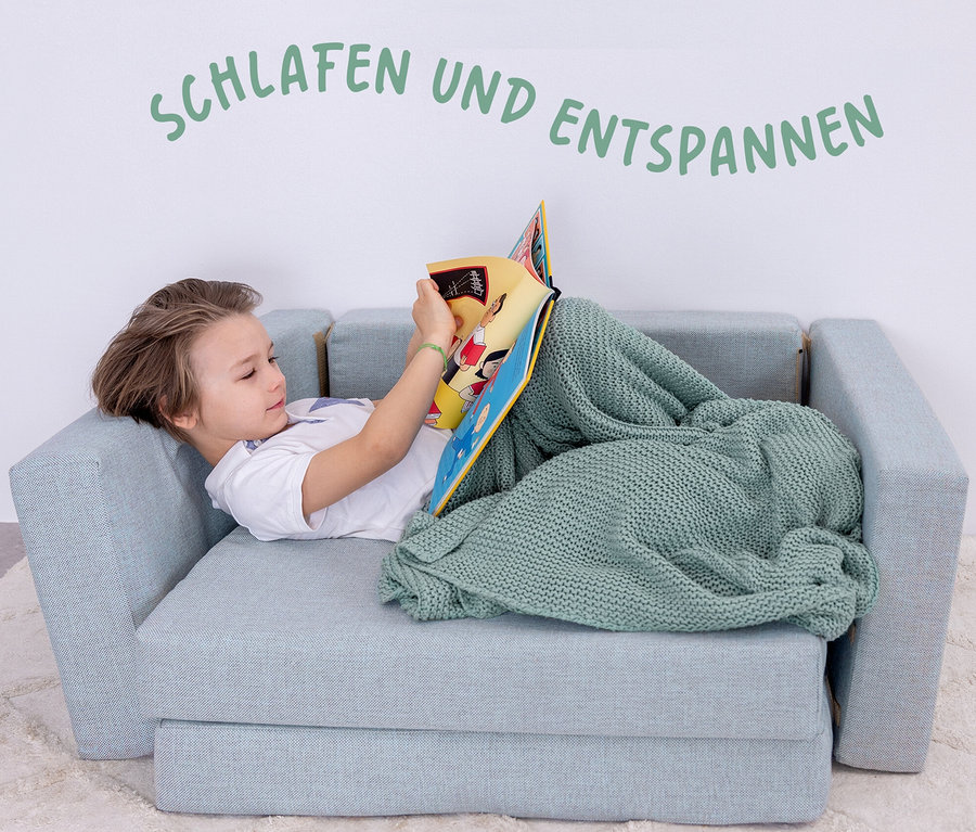 Junge liegt auf mintgrauem Buddies Spielsofa und liest ein Buch, zugedeckt mit einer grünen Decke.