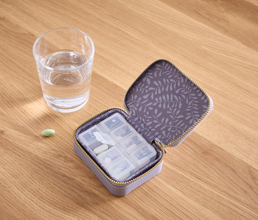 Auf einem Holztisch stehen ein Glas Wasser, eine grüne Pille und eine geöffnete violette Pillendose mit weißen Pillen darin.