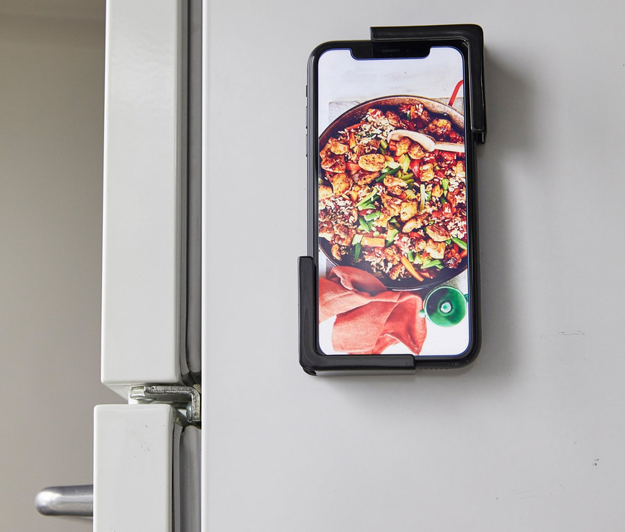 Ein Handy mit einem Bild von Essen ist mit einem schwarzen YAMAZAKI TOWER Tablethalter an einem weißen Kühlschrank befestigt.