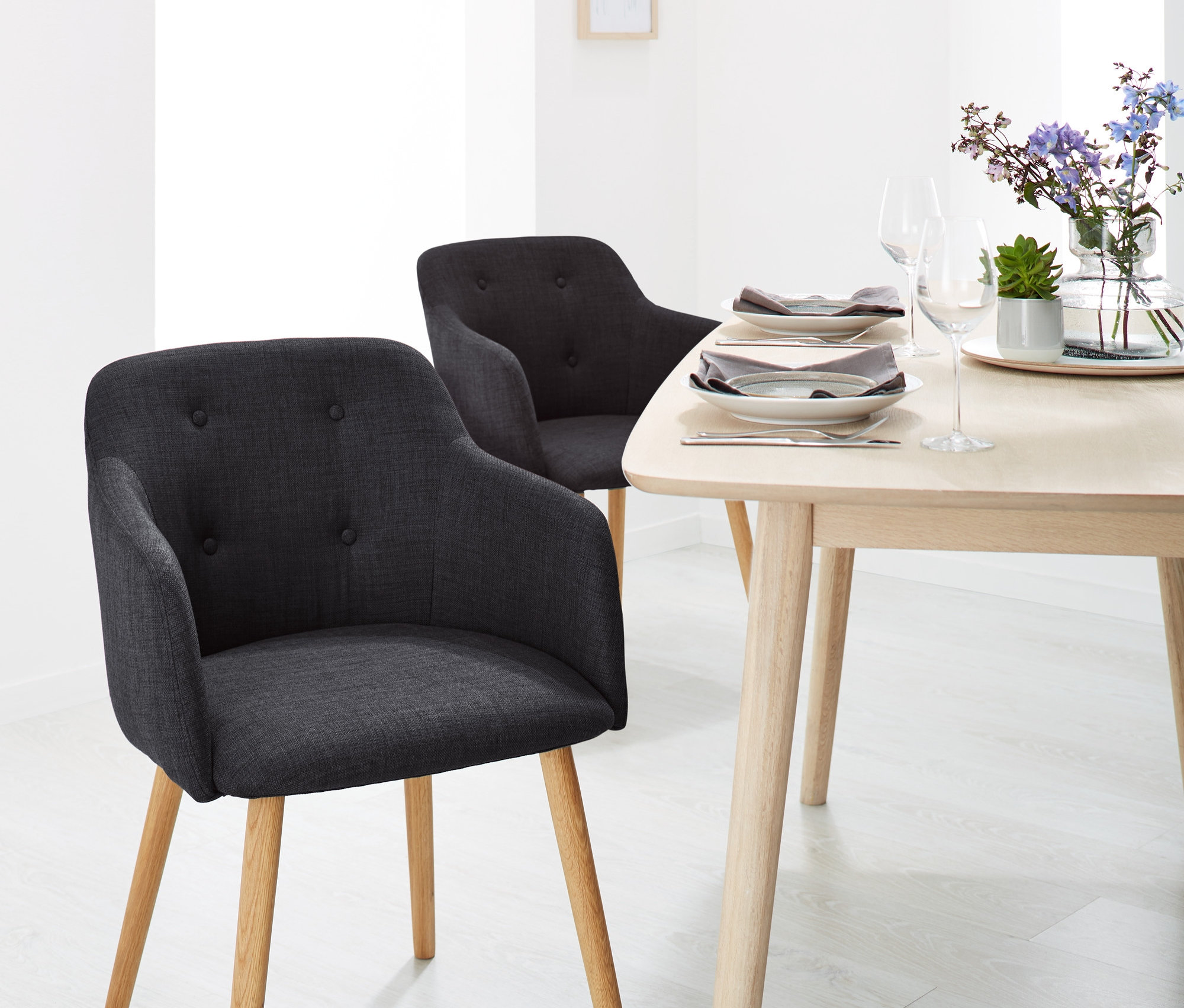 Zwei anthrazitfarbene, gepolsterte Stühle mit Armlehnen und ein Holztisch mit Blumen und Tellern in einem Esszimmer.