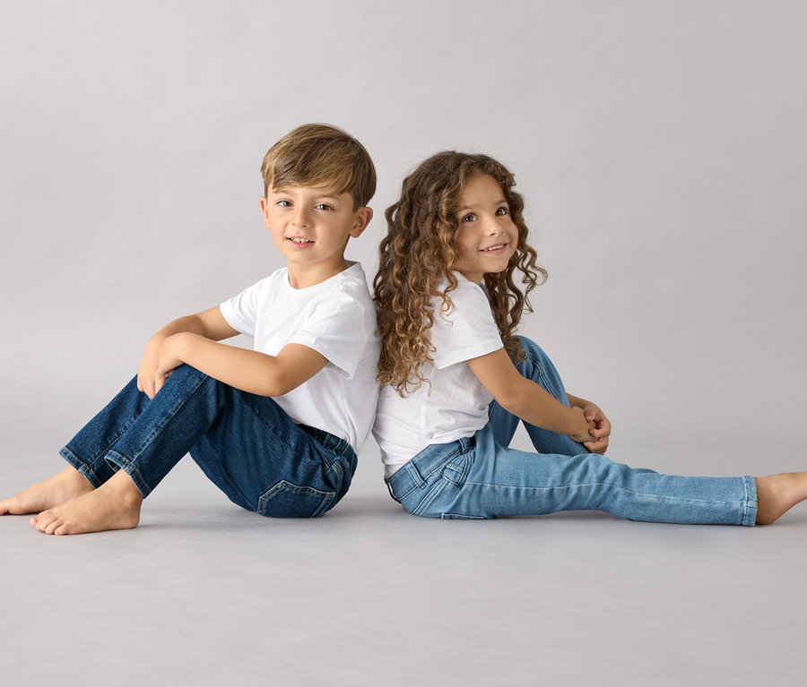 Ein Junge und ein Mädchen sitzen Rücken an Rücken und tragen beide weiße T-Shirts und Kinder-Pull-on-Pants – Fit »Luca«.