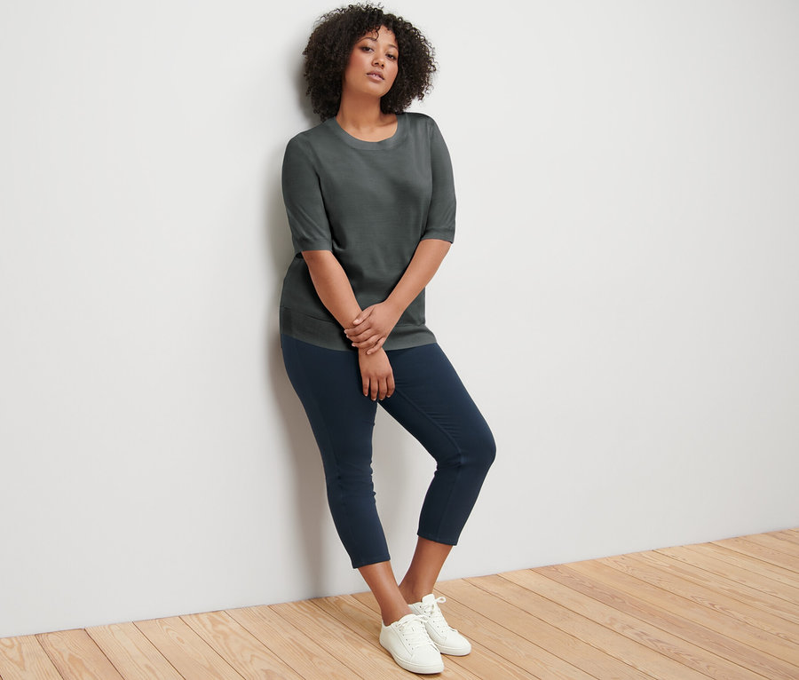 Eine Frau lehnt an einer Wand und trägt einen grünen Feinstrickpullover, dunkelblaue Hosen und weiße Turnschuhe.