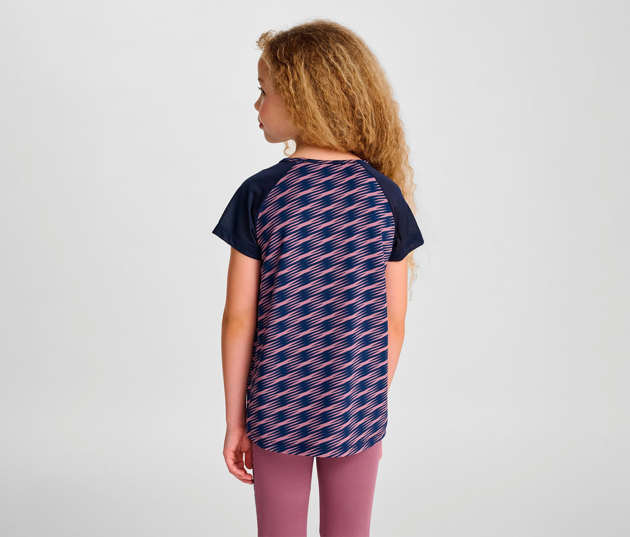 Mädchen steht mit dem Rücken zur Kamera und trägt ein HUMMEL HMLJR Mix T-Shirt S/S und rosa Leggings.