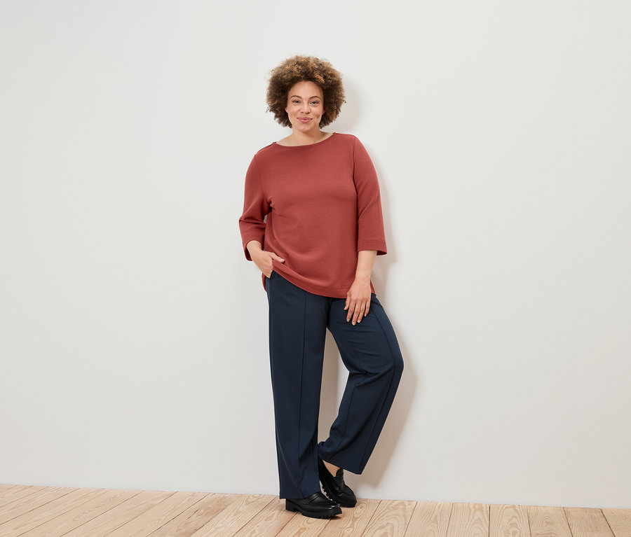 Frau mit lockigem Haar steht in brauner Bluse und dunkelblauer Hose an einer Wand.