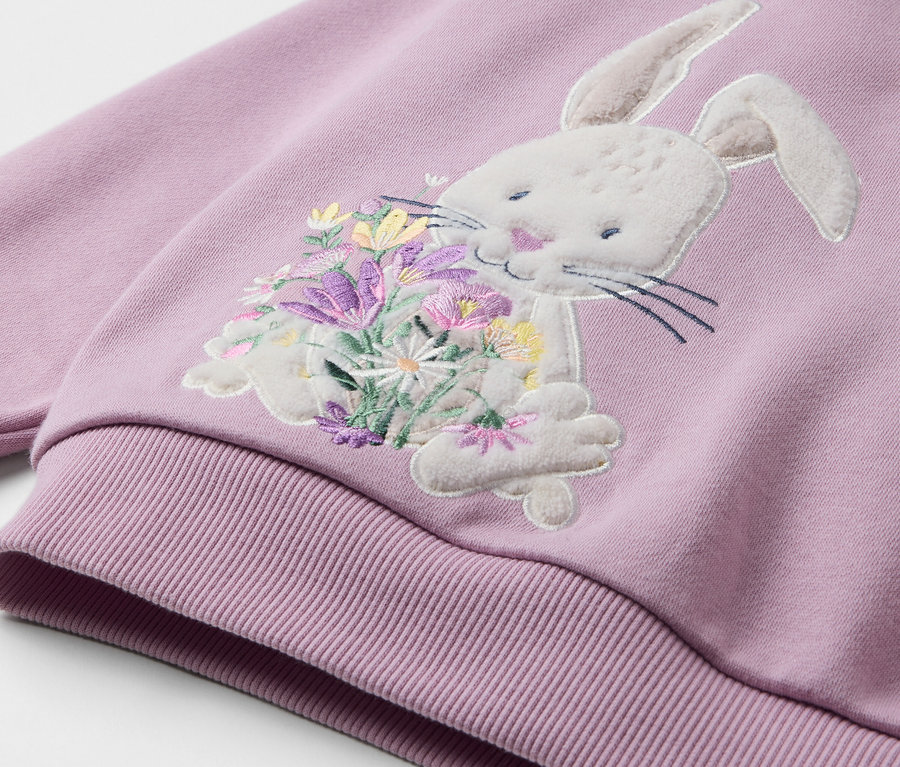 Detailansicht eines helllila Sweatshirts mit einer Applikation eines Hasen, der einen Blumenstrauß hält.