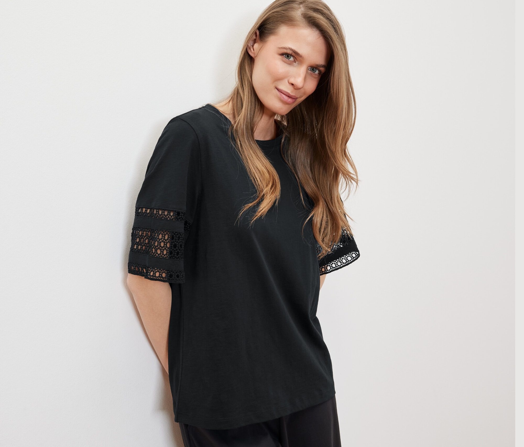 Eine Frau, die sich an eine Wand lehnt, trägt ein schwarzes Shirt mit Stickerei.