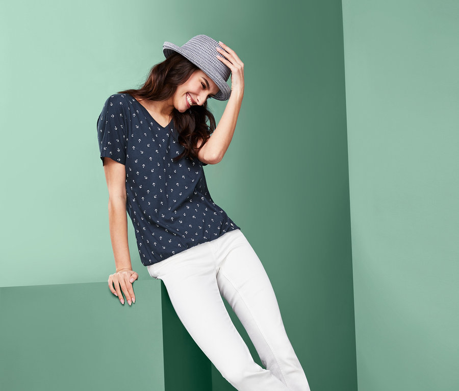 Eine Frau sitzt auf der Kante eines grünen Würfels und lächelt. Sie trägt ein Blusenshirt, eine weisse Hose und Turnschuhe. Auf dem Kopf hat sie einen Hut.