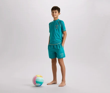 Ein Junge steht barfuß mit einem türkisfarbenen T-Shirt und Shorts neben einem bunten Ball vor einem grauen Hintergrund.