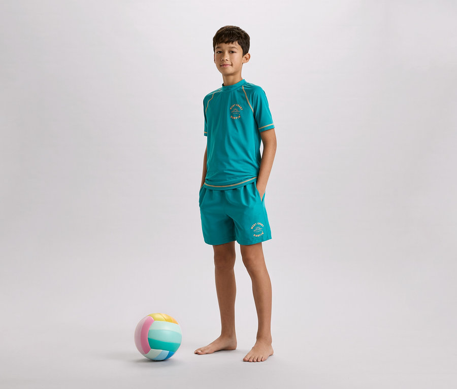 Ein Junge steht barfuß mit einem türkisfarbenen T-Shirt und Shorts neben einem bunten Ball vor einem grauen Hintergrund.