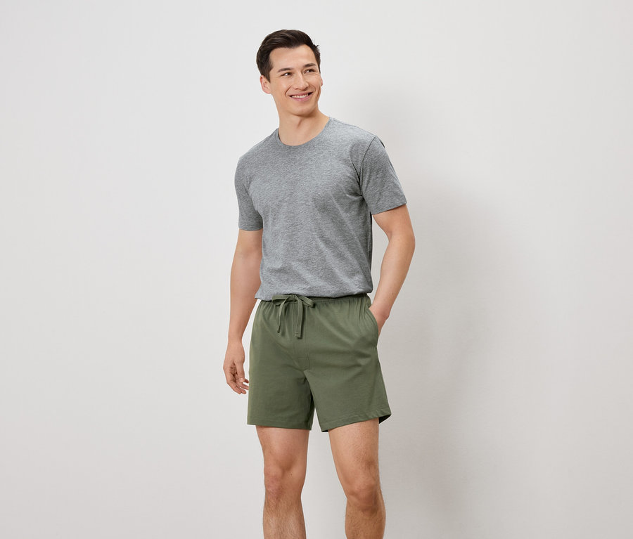 Mann mit grauem T-Shirt und grünen Shorts steht vor einem weißen Hintergrund.