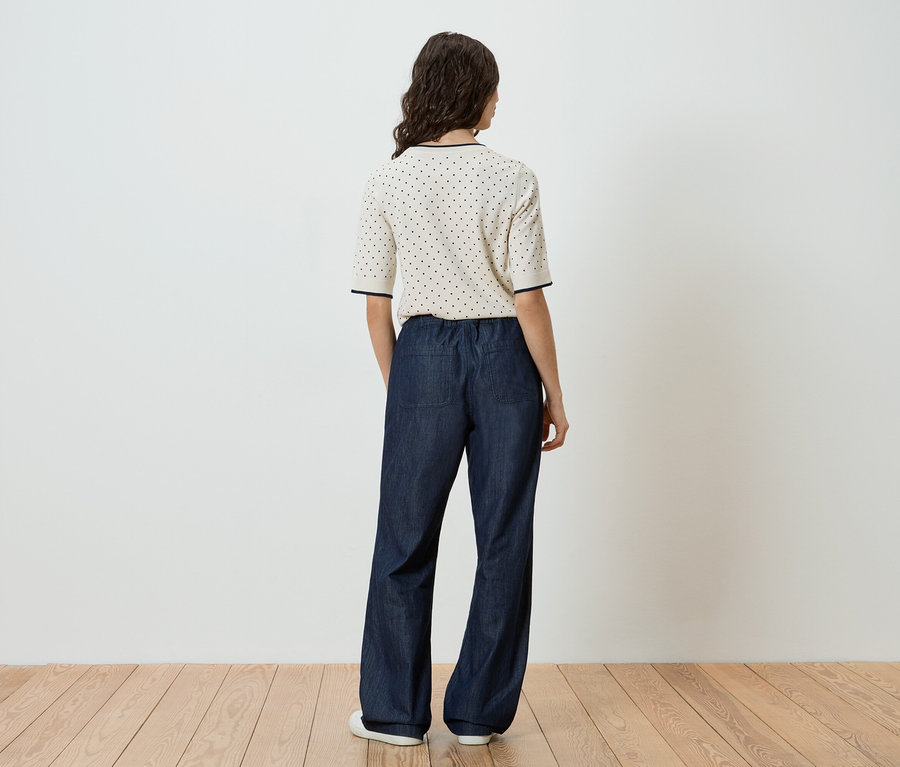 Rückansicht einer Frau mit dunklen, gewellten Haaren, die ein kurzärmeliges, cremefarbenes Oberteil mit marineblauen Punkten und eine weite, dunkelblaue Hose mit elastischem Bund und weißen Schuhen trägt.