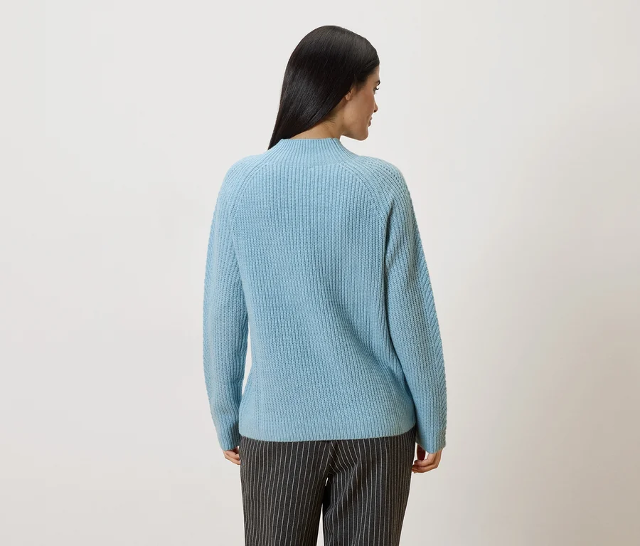 Rückansicht einer Frau mit langen, dunklen Haaren, die einen hellblauen Pullover und eine grau gestreifte Hose trägt.