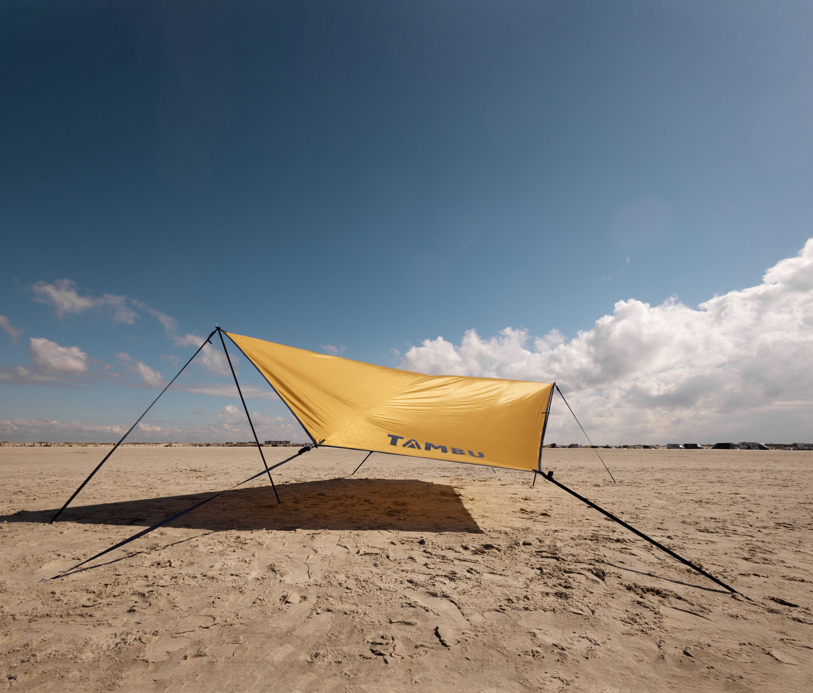 Ein gelbes Tambu »PALA« Tarp/Sonnensegel steht am Strand unter blauem Himmel.