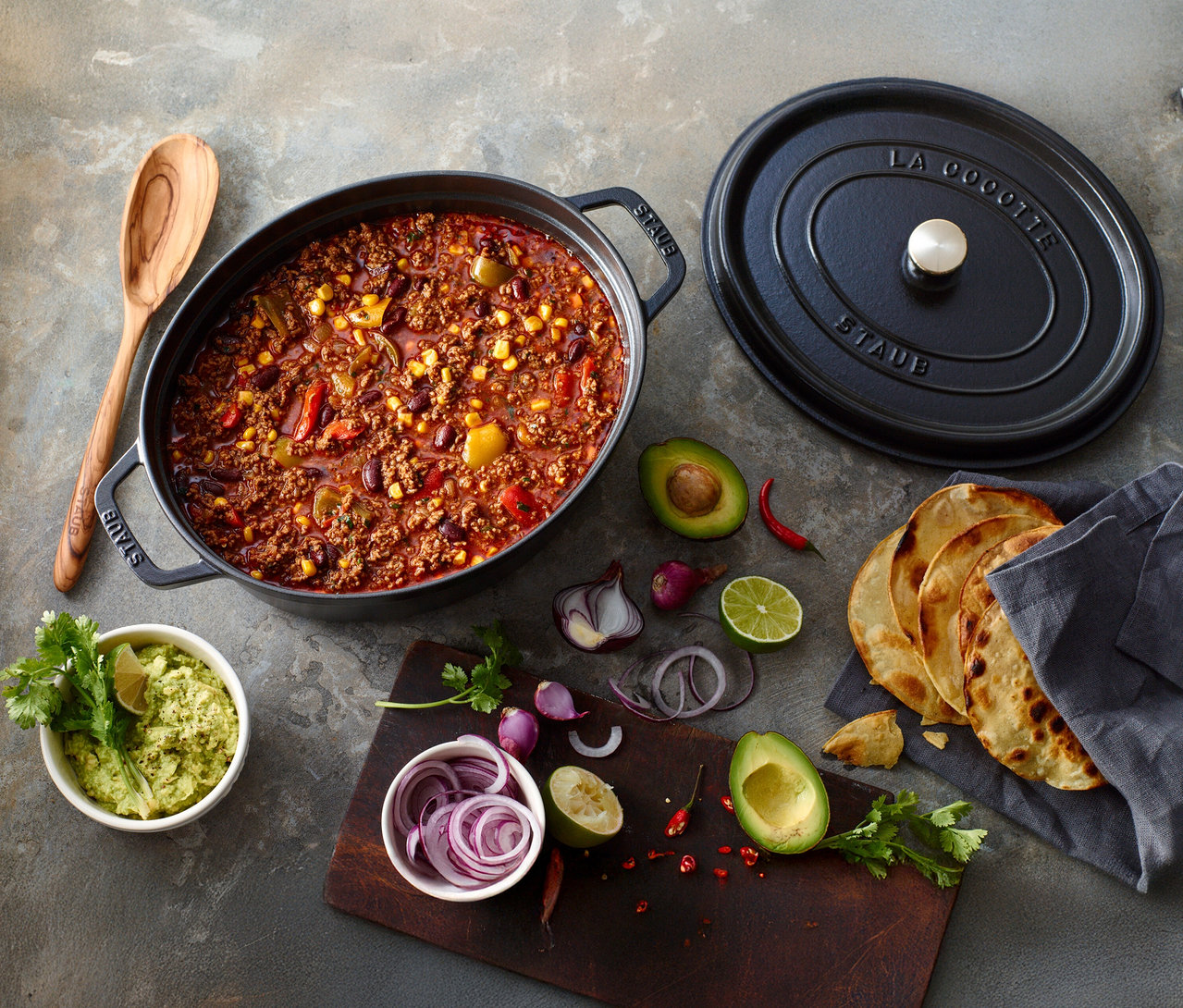 Chili con Carne im schwarzen STAUB Bräter »Cocotte 33« (ca. 6,7 l) mit Zutaten wie Avocado, Zwiebeln und Tortillas.