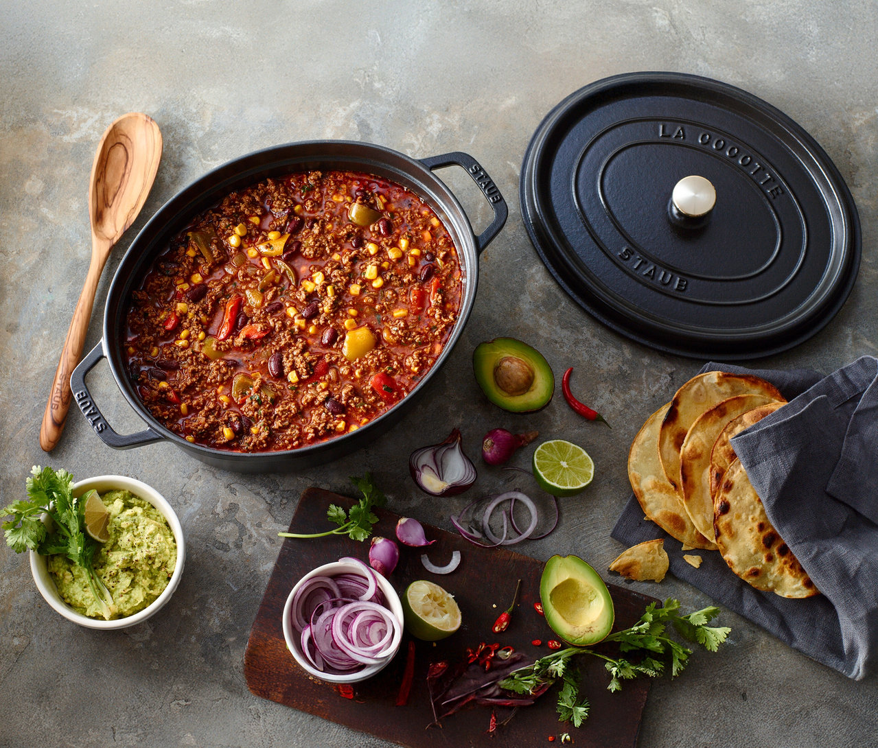 Chili con Carne im schwarzen STAUB Bräter »Cocotte 29« mit Holzlöffel, Guacamole, Zwiebeln, Limetten und Tortillas.