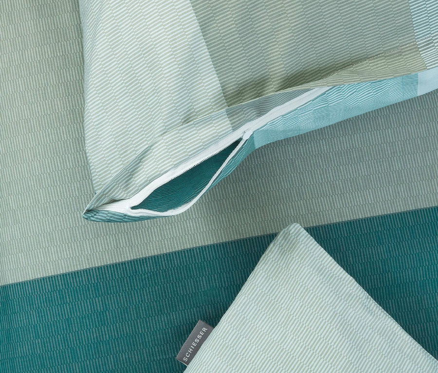 Nahaufnahme von Schiesser-Bettwäsche mit geometrischen Streifen in Grün- und Blautönen, teilweise geöffnetem Reißverschluss und einem Kissen mit dem Schiesser-Logo.