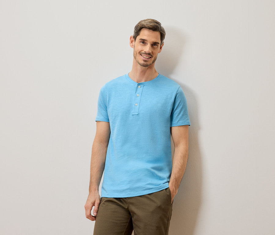 Ein Mann mit kurzen braunen Haaren und Bart, der ein hellblaues T-Shirt mit Knöpfen und eine Khakihose trägt, steht vor einer hellgrauen Wand.