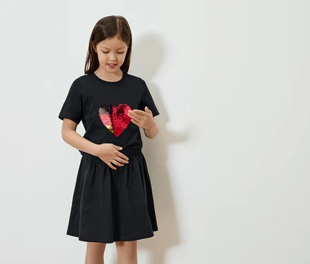 Mädchen in schwarzem T-Shirt mit Wendepailletten-Herz und Rock posiert vor einer weißen Wand.