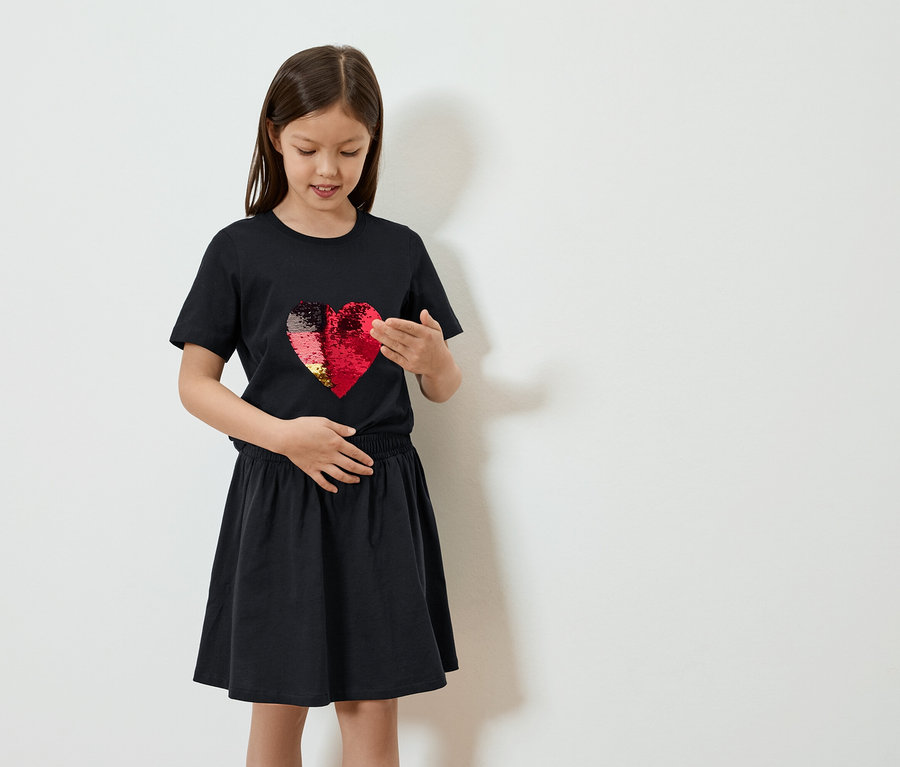 Mädchen in schwarzem T-Shirt mit Wendepailletten-Herz und Rock posiert vor einer weißen Wand.