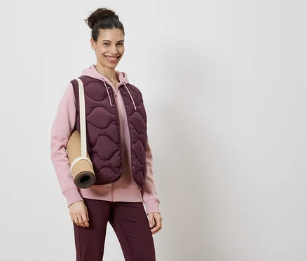 Frau trägt eine burgunderfarbene Steppweste über einem rosa Kapuzenpullover und burgunderfarbene Leggings, mit einer Yogamatte über der Schulter.