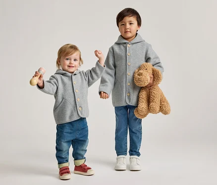 Ein Kleinkind und ein älteres Kind stehen nebeneinander. Beide tragen graue Kapuzenjacken mit Holzknöpfen und blaue Jeans. Das Kleinkind links hält eine Holzrassel und trägt rote Schuhe. Das ältere Kind rechts hält einen braunen Plüschhund und trägt weiße Schuhe.