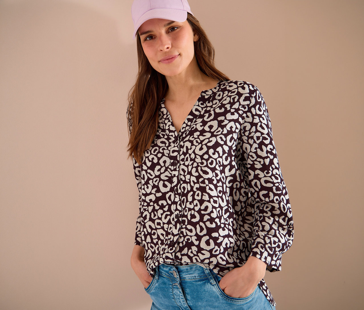 Frau mit rosa Kappe und gemusterter Bluse, die ihre Hände in die Jeanshosentaschen steckt.
