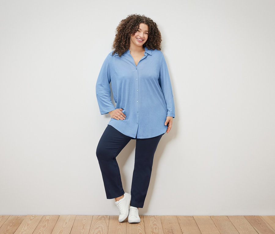 Frau mit lockigem Haar trägt eine hellblaue Bluse, marineblaue Hose und weiße Schuhe.