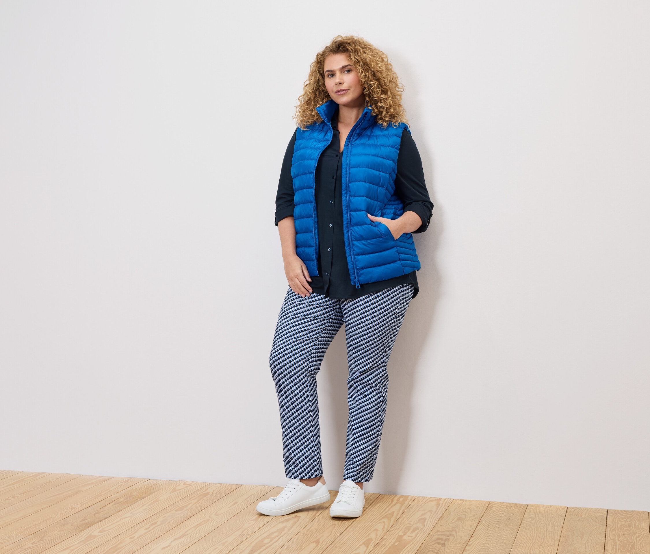 Frau mit lockigem Haar, blauer Weste, dunklem Hemd und gemusterten Hosen steht an einer weißen Wand.
