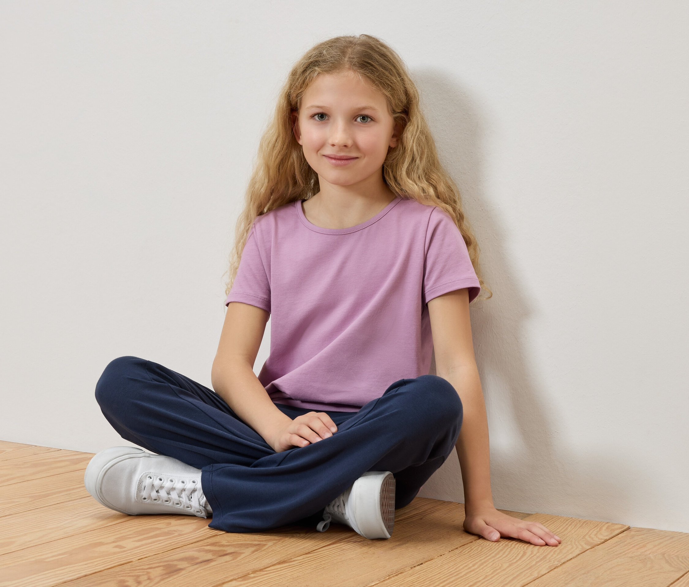Mädchen mit blonden Haaren sitzt mit gekreuzten Beinen auf einem Holzboden, ein lila T-Shirt und blaue Hosen tragend.
