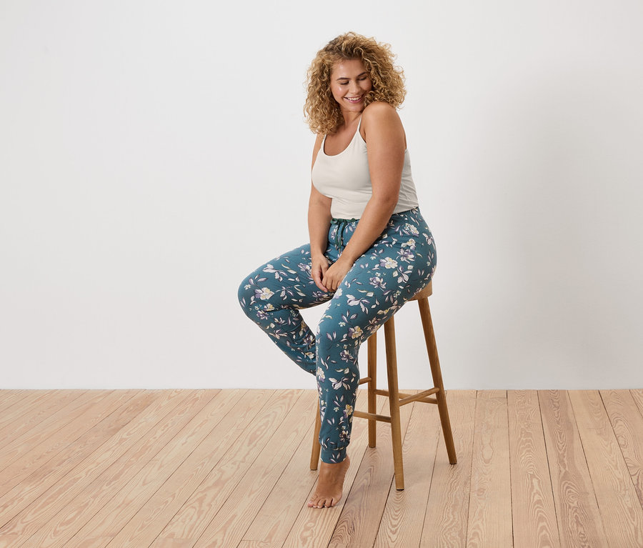 Frau mit lockigem Haar sitzt auf einem Hocker und trägt ein Tanktop und blumige Leggings.