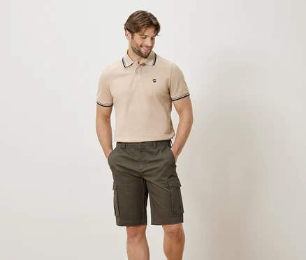 Mann mit beigefarbenem Poloshirt und olivgrünen Cargoshorts posiert mit Händen in den Hosentaschen vor einem weißen Hintergrund.