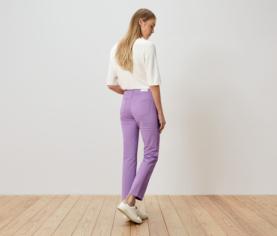 Frau von hinten, mit lila Hose und weißem Oberteil, steht auf einem Holzboden vor einer weißen Wand.