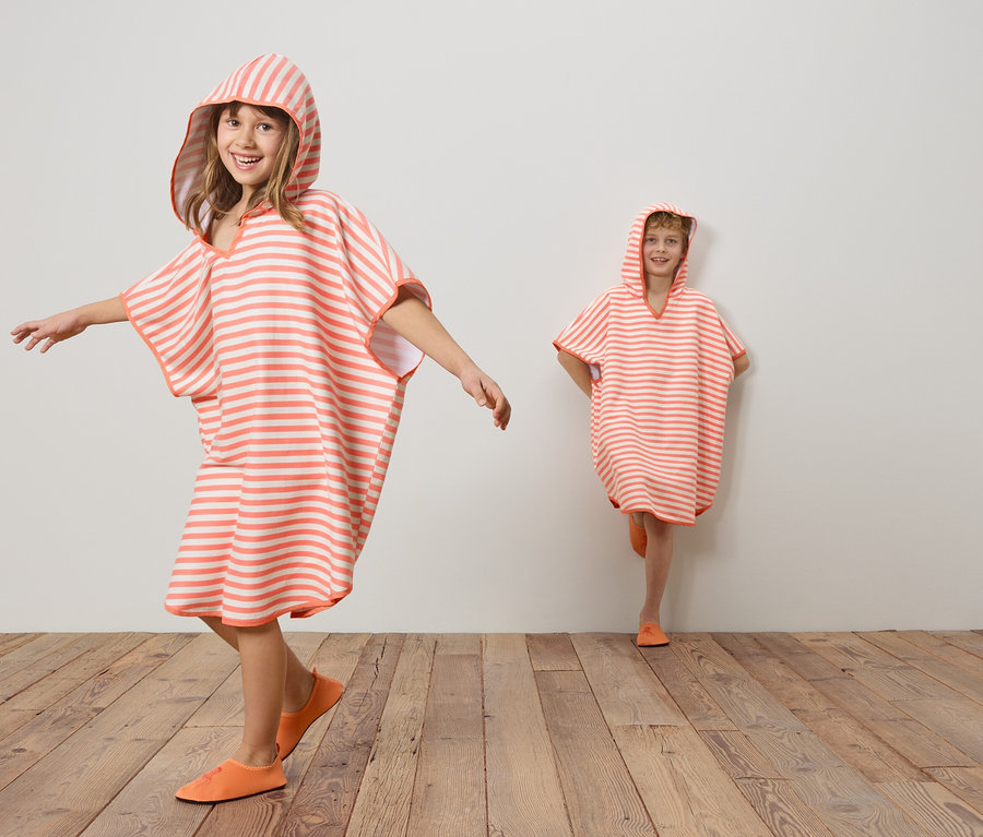 Zwei Kinder tragen gestreifte, korallenrote und weiße Kapuzenponchos und korallenrote Schuhe auf einem Holzboden vor einer weißen Wand.