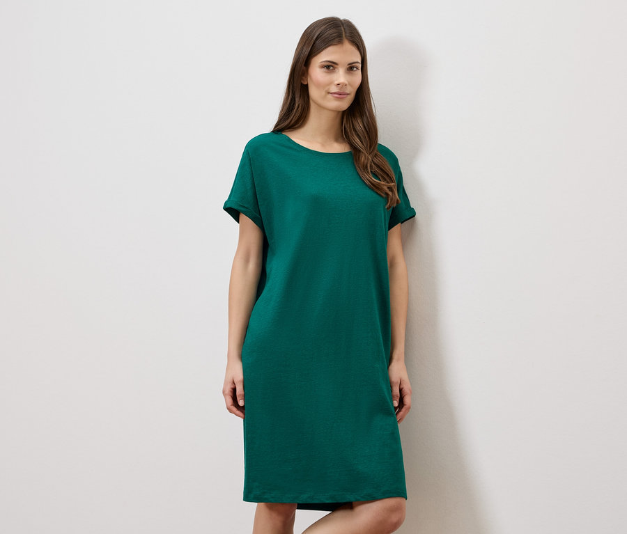 Frau trägt ein grünes, kurzes Kleid vor einer weißen Wand.